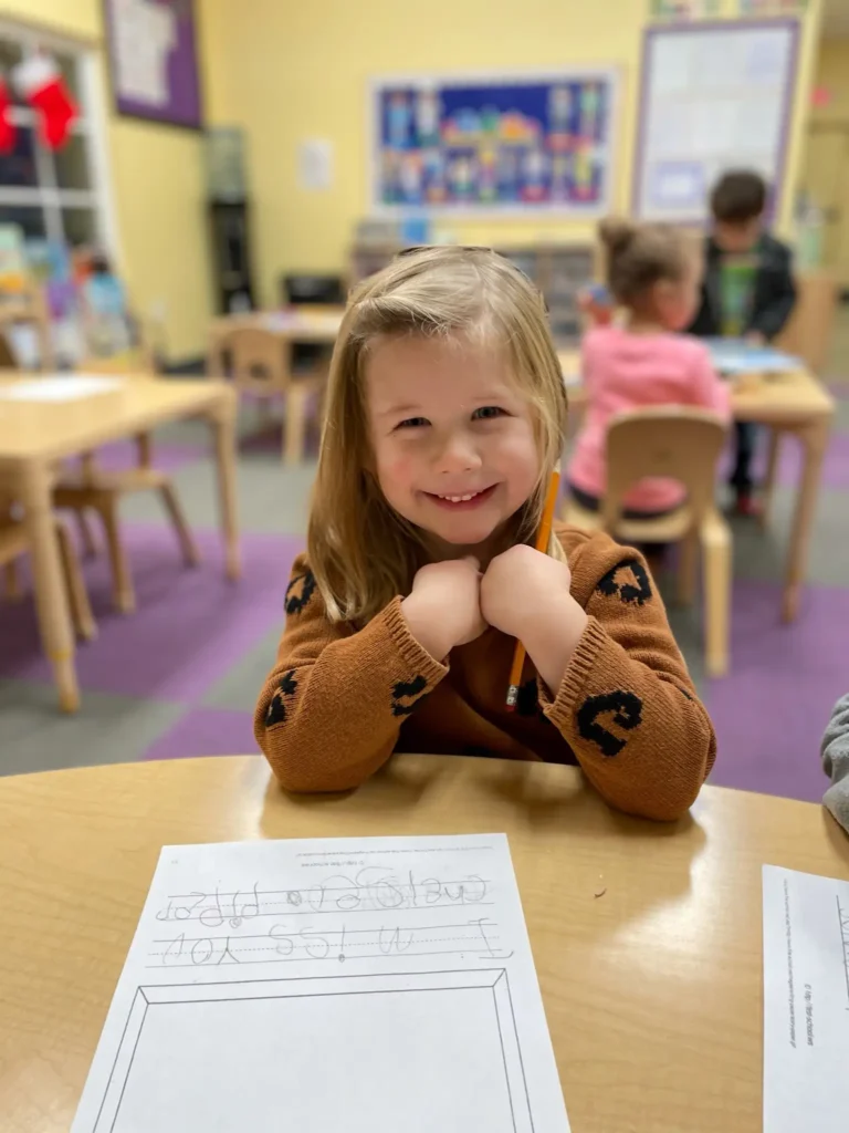 Girl smiling at a table while working on a preschool writing activity.